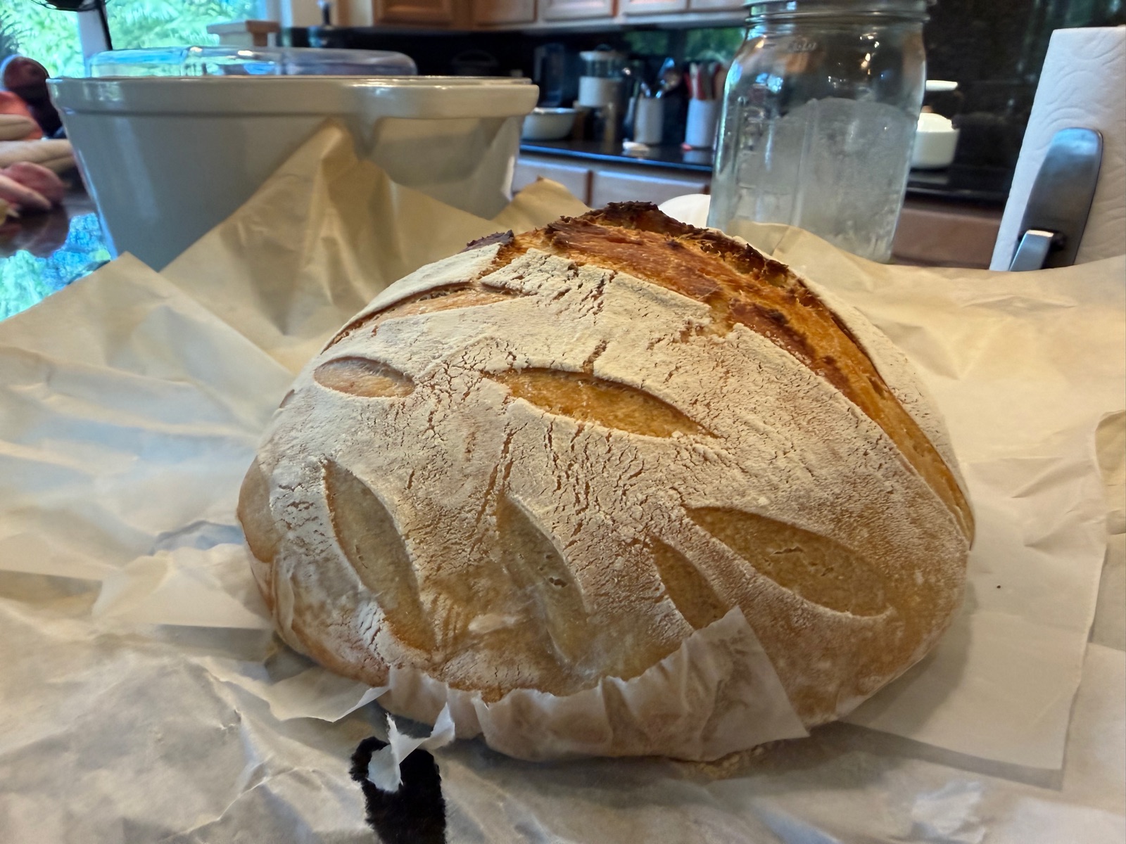 A scored sourdough boule on parchment, with the Wahine starter jar in the background.